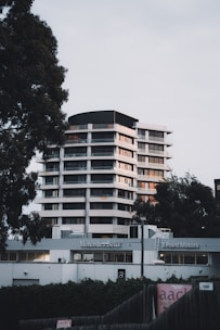 A modern high-rise building is located behind a Mercedes-Benz dealership, flanked by tall trees on either side. The structure features multiple floors with a sleek design, utilizing large windows and white exterior paneling. The foreground includes dealership signage and a fence partially covered with ivy.