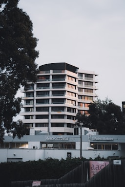 A modern high-rise building is located behind a Mercedes-Benz dealership, flanked by tall trees on either side. The structure features multiple floors with a sleek design, utilizing large windows and white exterior paneling. The foreground includes dealership signage and a fence partially covered with ivy.