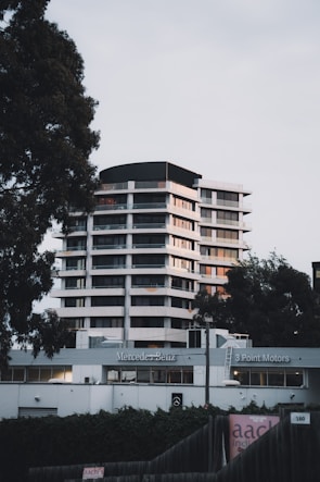 A modern high-rise building is located behind a Mercedes-Benz dealership, flanked by tall trees on either side. The structure features multiple floors with a sleek design, utilizing large windows and white exterior paneling. The foreground includes dealership signage and a fence partially covered with ivy.