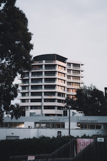 A modern high-rise building is located behind a Mercedes-Benz dealership, flanked by tall trees on either side. The structure features multiple floors with a sleek design, utilizing large windows and white exterior paneling. The foreground includes dealership signage and a fence partially covered with ivy.