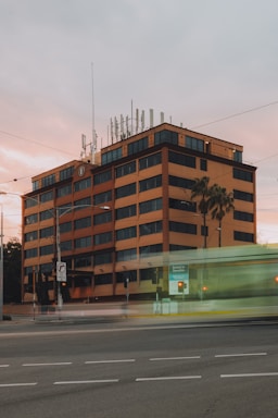 A modern, multi-story office building with a reddish-brown facade and numerous windows is located along a busy street. The building is equipped with several antennas on the roof. A tram is visible in motion, creating a blur effect on the lower part of the image. The sky has a soft, pastel tone suggesting either sunrise or sunset.