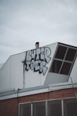 A section of a building rooftop features a graffiti piece with bold, stylized letters. The structure includes a white panel with an angled design element, and there's a chimney pipe protruding near the graffiti. Below the graffiti, part of the building façade is visible, showcasing a brick wall and metal mesh.