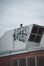 A section of a building rooftop features a graffiti piece with bold, stylized letters. The structure includes a white panel with an angled design element, and there's a chimney pipe protruding near the graffiti. Below the graffiti, part of the building façade is visible, showcasing a brick wall and metal mesh.