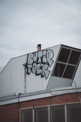 A section of a building rooftop features a graffiti piece with bold, stylized letters. The structure includes a white panel with an angled design element, and there's a chimney pipe protruding near the graffiti. Below the graffiti, part of the building façade is visible, showcasing a brick wall and metal mesh.