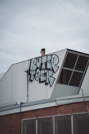 A section of a building rooftop features a graffiti piece with bold, stylized letters. The structure includes a white panel with an angled design element, and there's a chimney pipe protruding near the graffiti. Below the graffiti, part of the building façade is visible, showcasing a brick wall and metal mesh.