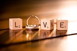 Four wooden blocks each displaying letters and numbers, arranged to spell the word 'LOVE'. A silver ring is intertwined with the letter 'O'. The scene is warmly lit, casting soft shadows on a wooden surface.
