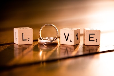 Four wooden blocks each displaying letters and numbers, arranged to spell the word 'LOVE'. A silver ring is intertwined with the letter 'O'. The scene is warmly lit, casting soft shadows on a wooden surface.