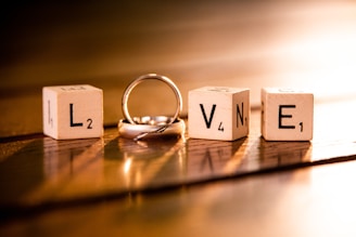 Four wooden blocks each displaying letters and numbers, arranged to spell the word 'LOVE'. A silver ring is intertwined with the letter 'O'. The scene is warmly lit, casting soft shadows on a wooden surface.