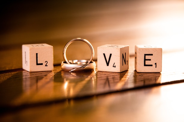 Four wooden blocks each displaying letters and numbers, arranged to spell the word 'LOVE'. A silver ring is intertwined with the letter 'O'. The scene is warmly lit, casting soft shadows on a wooden surface.
