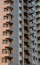 A high-rise building featuring a series of balconies with air conditioning units on each floor and a side view of an external staircase running vertically. The structure exhibits a modern architectural style with a mix of beige and gray tones.