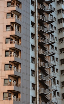 A high-rise building featuring a series of balconies with air conditioning units on each floor and a side view of an external staircase running vertically. The structure exhibits a modern architectural style with a mix of beige and gray tones.