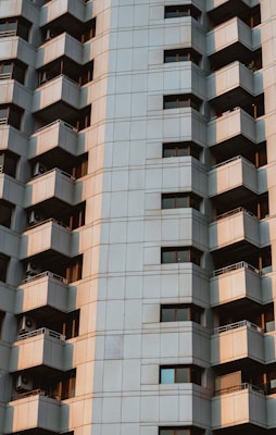 A tall residential building with a modern design features numerous balconies on each floor. The facade comprises a grid of square panels in a muted beige tone, and the balconies include metal railings. The building has several windows reflecting sunlight, adding a warm tone to the scene.