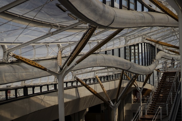A complex structure composed of curving metal tubes and supports, integrated into a building with large glass windows and an industrial design. The architecture includes staircases, creating a sense of movement and fluidity amid the steel and glass materials.