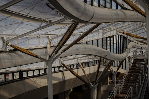 A complex structure composed of curving metal tubes and supports, integrated into a building with large glass windows and an industrial design. The architecture includes staircases, creating a sense of movement and fluidity amid the steel and glass materials.