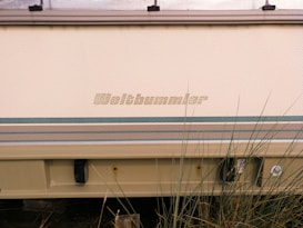 A close-up view of the lower part of a vehicle featuring the name 'Weltbummler' on its side. The vehicle has a textured beige surface with striped patterns in shades of green and blue. The lower section includes a beige bumper and some exposed screws or fixtures. Tall grass partially obscures the bottom of the image.