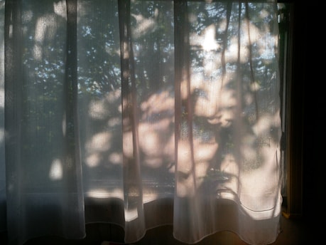 Sheer curtains hang in front of a window, with sunlight casting leafy patterns and shadows onto the fabric. The light filtering through creates a soft and serene atmosphere.