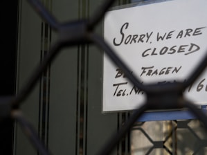 A sign with handwritten text in English and German is visible through a metal gate or grille. The sign reads 'Sorry, we are closed,' suggesting the closure of a business or shop.