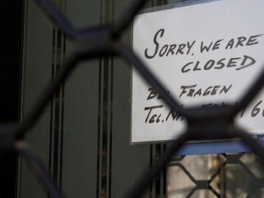 A sign with handwritten text in English and German is visible through a metal gate or grille. The sign reads 'Sorry, we are closed,' suggesting the closure of a business or shop.