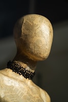 A mannequin head made from paper mache is adorned with pages of text. It has a paper-like texture and wears a black lace choker. The lighting casts soft shadows, highlighting the contours of the head and neck.