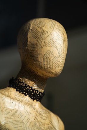 A mannequin head made from paper mache is adorned with pages of text. It has a paper-like texture and wears a black lace choker. The lighting casts soft shadows, highlighting the contours of the head and neck.