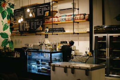 A cozy cafe interior with a barista preparing drinks behind the counter. Shelves lined with various packaged goods and cups are prominently displayed. A menu board with illuminated bulbs hangs above the counter. The scene includes a refrigerator and a freezer while leafy green wall decorations add a touch of nature.