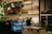 A cozy cafe interior with a barista preparing drinks behind the counter. Shelves lined with various packaged goods and cups are prominently displayed. A menu board with illuminated bulbs hangs above the counter. The scene includes a refrigerator and a freezer while leafy green wall decorations add a touch of nature.