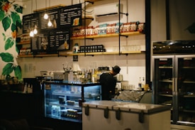A cozy cafe interior with a barista preparing drinks behind the counter. Shelves lined with various packaged goods and cups are prominently displayed. A menu board with illuminated bulbs hangs above the counter. The scene includes a refrigerator and a freezer while leafy green wall decorations add a touch of nature.