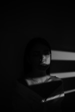 A person stands in a dimly lit environment with shadows and light creating dramatic patterns across their face and torso. The mood is introspective and somber, with the play of light highlighting the contours of the face.