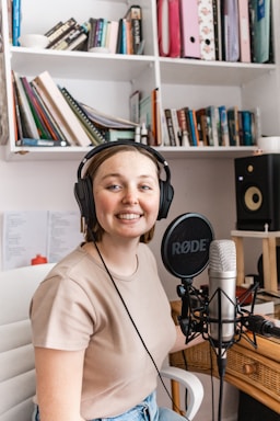 A person wearing headphones is sitting in front of a microphone in a room with bookshelves filled with various books and files. The person is smiling, suggesting a friendly atmosphere. There is also a speaker on one of the shelves.