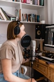 A person wearing headphones is sitting at a desk with a microphone in front of them, presumably recording or working with audio. There is a computer screen displaying audio software, and several books and folders are neatly arranged on shelves behind.