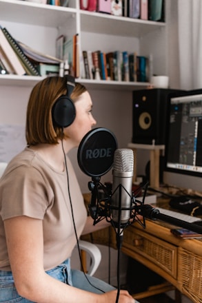 A person wearing headphones is sitting at a desk with a microphone in front of them, presumably recording or working with audio. There is a computer screen displaying audio software, and several books and folders are neatly arranged on shelves behind.