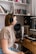 A person wearing headphones is sitting at a desk with a microphone in front of them, presumably recording or working with audio. There is a computer screen displaying audio software, and several books and folders are neatly arranged on shelves behind.