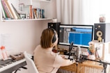 A person is sitting at a desk working on music production. They are wearing headphones and facing a large computer screen displaying a digital audio workstation interface. Surrounding them are various items such as a microphone on a stand, studio speakers, a keyboard, books on shelves, and a water bottle.