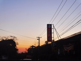 A serene evening scene with a clear gradient in the sky transitioning from light blue to warm orange hues. Silhouettes of trees and power lines stretch across the image, with a prominent sign featuring red text and a logo. The lower part of the image contains shadows of buildings and a visible parking sign with a 'P'.