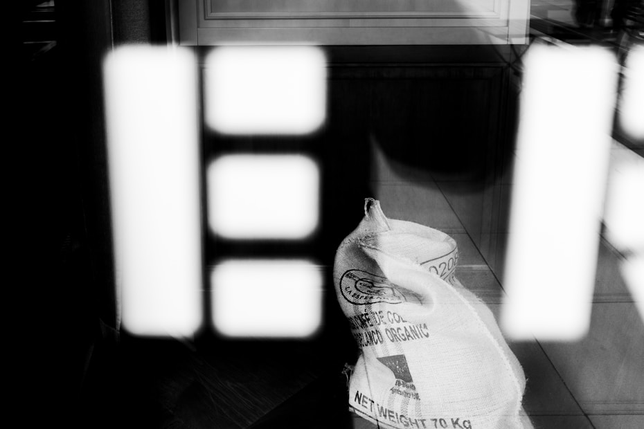 A burlap sack labeled 'Cultivado Organico' and 'Net Weight 70 KG' is placed against a reflective surface that shows abstract, blurred reflections of geometric shapes in high contrast black and white.