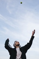 A person is preparing to hit a tennis ball with a racket. They have their eyes on the ball which is high in the air. The person is wearing a white headband, a white shirt, and a dark jacket. The sky above is clear with a few wispy clouds.