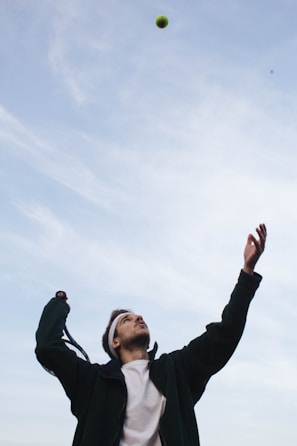 A person is preparing to hit a tennis ball with a racket. They have their eyes on the ball which is high in the air. The person is wearing a white headband, a white shirt, and a dark jacket. The sky above is clear with a few wispy clouds.