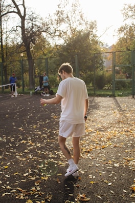 A person wearing white sportswear is standing on a leaf-covered tennis court, illuminated by warm sunlight. In the background, other people are present near a fence, with trees showcasing autumn foliage partially obstructing the view.