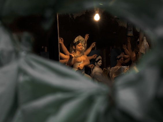 A dimly lit space showcases several clay sculptures of human figures, with a single light bulb hanging above, casting a warm glow. Some figures are wrapped in transparent plastic, and the scene is viewed through leaves or an opening, creating a sense of depth and intrigue.