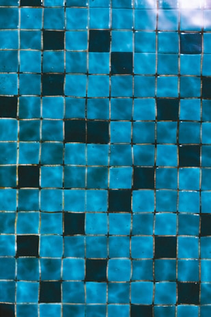 A grid of small, square blue tiles with a shiny, reflective surface. The tiles vary slightly in hue, creating a wave-like pattern due to lighting and reflections.