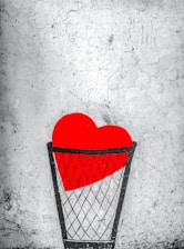 A bright red heart is placed inside a black wire basket against a textured, gray background. The heart is prominent and vibrant, contrasting with the muted and worn appearance of the wall.
