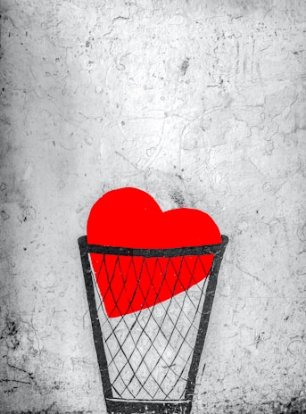 A bright red heart is placed inside a black wire basket against a textured, gray background. The heart is prominent and vibrant, contrasting with the muted and worn appearance of the wall.