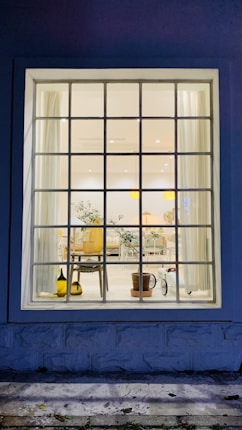 A large window showcases an interior view featuring a well-lit room with a mix of modern and natural decor. The room contains elements such as a potted plant, a wooden chair, and a side table. Light curtains frame the window, and soft lighting enhances the cozy ambiance. The floor appears clean and is complemented by simple, elegant furniture.
