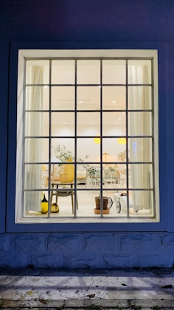 A large window showcases an interior view featuring a well-lit room with a mix of modern and natural decor. The room contains elements such as a potted plant, a wooden chair, and a side table. Light curtains frame the window, and soft lighting enhances the cozy ambiance. The floor appears clean and is complemented by simple, elegant furniture.