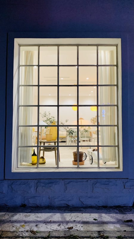 A large window showcases an interior view featuring a well-lit room with a mix of modern and natural decor. The room contains elements such as a potted plant, a wooden chair, and a side table. Light curtains frame the window, and soft lighting enhances the cozy ambiance. The floor appears clean and is complemented by simple, elegant furniture.