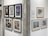 A gallery wall featuring a collection of framed artworks. On the left, six pieces are displayed in black frames, showcasing various abstract and figurative styles. On the right, three pieces in lighter frames, with themes ranging from abstraction to realistic imagery. A textured column separates the two sections, and a rectangular plaque provides information about the display.
