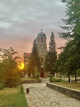 A stone pathway leads to a large church with a prominent dome and a cross on top, surrounded by tall evergreen trees. The sky is filled with soft, colorful clouds at sunset, casting a warm glow across the scene. Lampposts line the path, adding a quaint and serene atmosphere.