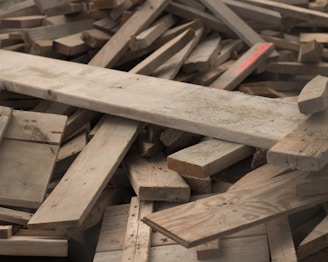 A pile of variously sized wooden planks stacked haphazardly. The wood appears weathered and includes a mix of light and dark tones with visible grain and knot patterns.