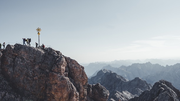 Several people hike along a rocky mountain peak, with a prominent golden cross marking the summit. The surrounding landscape showcases expansive mountain ranges under a clear, pale sky.