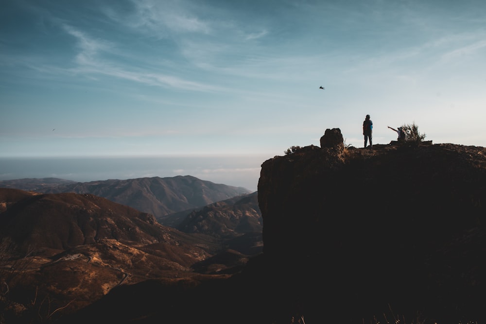 silhouette of 2 people standing on rock formation during daytime